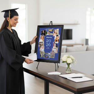 A smiling woman in a graduation gown stands next to a framed collage of graduation photos, with a vase of white flowers and papers on a table in front of her.
